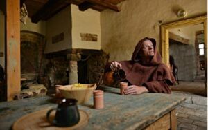 Středověký skotský mnich - sedí za stolem a nalévá si do sklenice whisky (nějaký alkoholický nápoj)