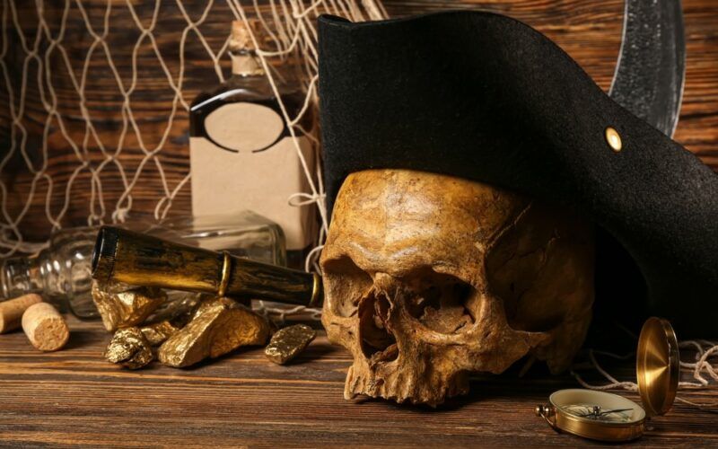 Piráti a rum: Lidská lebka s pirátským kloboukem, zlatými nugetkami, lahví rumu a cestovním vybavením na hnědém dřevě